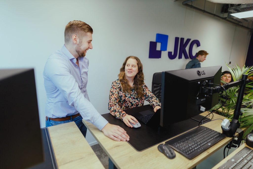 Two colleagues talking at a workstation under the JKC logo in the office